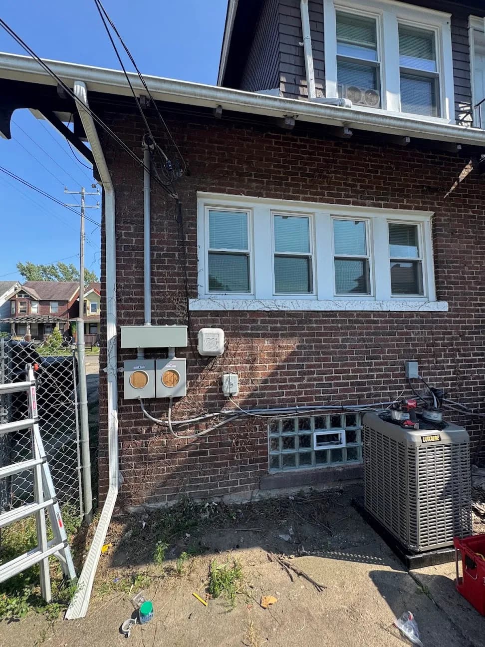 An electrical meter and service equipment on the brick exterior of a house, beside an air conditioning condenser
